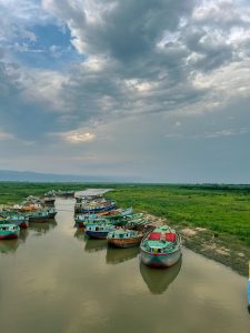 A serene view of a river lined with colorful fishing boats, moored at various angles. The boats are painted in shades of green, red, and blue, some adorned with decorative designs. Lush green vegetation surrounds the waterway, while distant hills and overcast skies add to the tranquil atmosphere. The water reflects the colors of the boats and the muted light from the sky, creating a harmonious natural scene.
