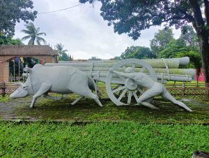 A white statue depicts a farmer straining to pull a wooden cart loaded with logs, which is being guided by a cow. The scene is set on a grassy area with tropical trees and colorful flags in the background, under a cloudy sky. The figures are stylized and represent traditional agricultural practices.