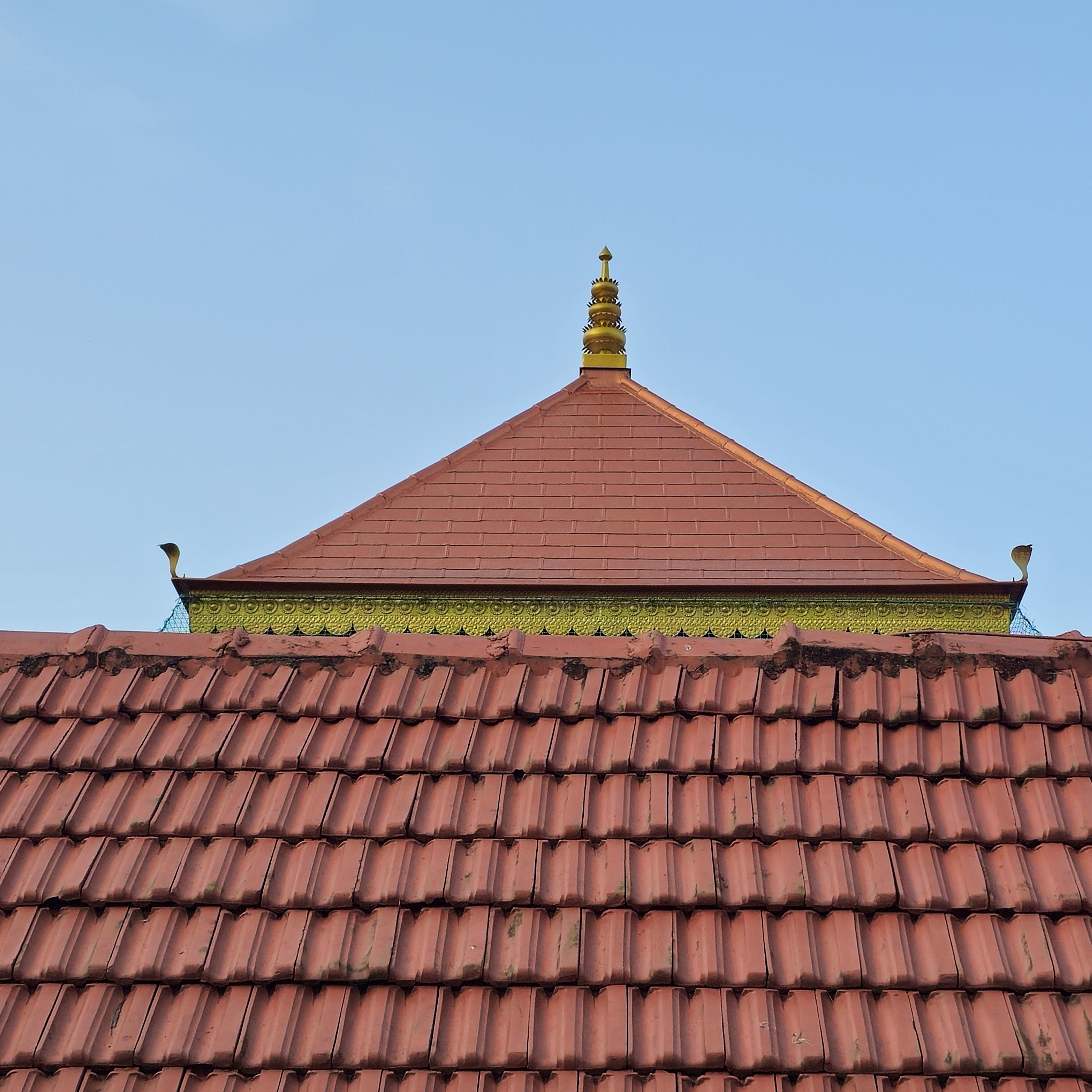 A traditional Kerala temple roof with red tiles and a golden finial stands bright under the blue sky. Captured from Gotheeswaram, Kozhikode.