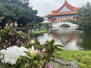 A tranquil scene in a garden, with a traditional East Asian-style building and a white arched bridge reflected in a pond. Pink and white flowers, likely azaleas, are in the foreground, with lush green trees and foliage surrounding the pond