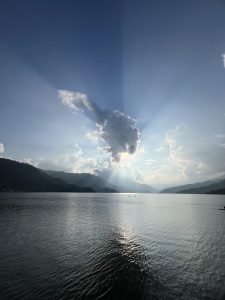 Sunlight streaming through dramatic clouds over a calm lake, surrounded by mountain silhouettes with two small boats visible in the distance.

