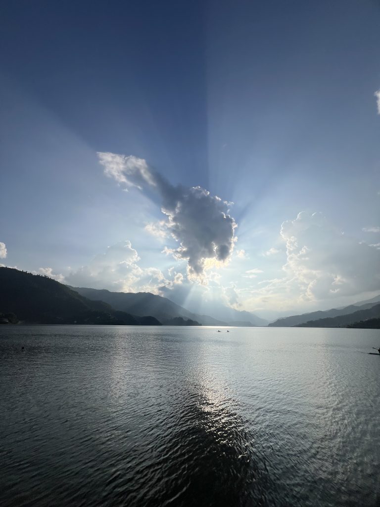 Sunlight streaming through dramatic clouds over a calm lake, surrounded by mountain silhouettes with two small boats visible in the distance.