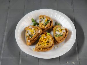  Four pieces of crispy Meetha Ghughra served on a white disposable plate, garnished with chopped onions, coriander leaves, and sev, placed on a grey surface.
