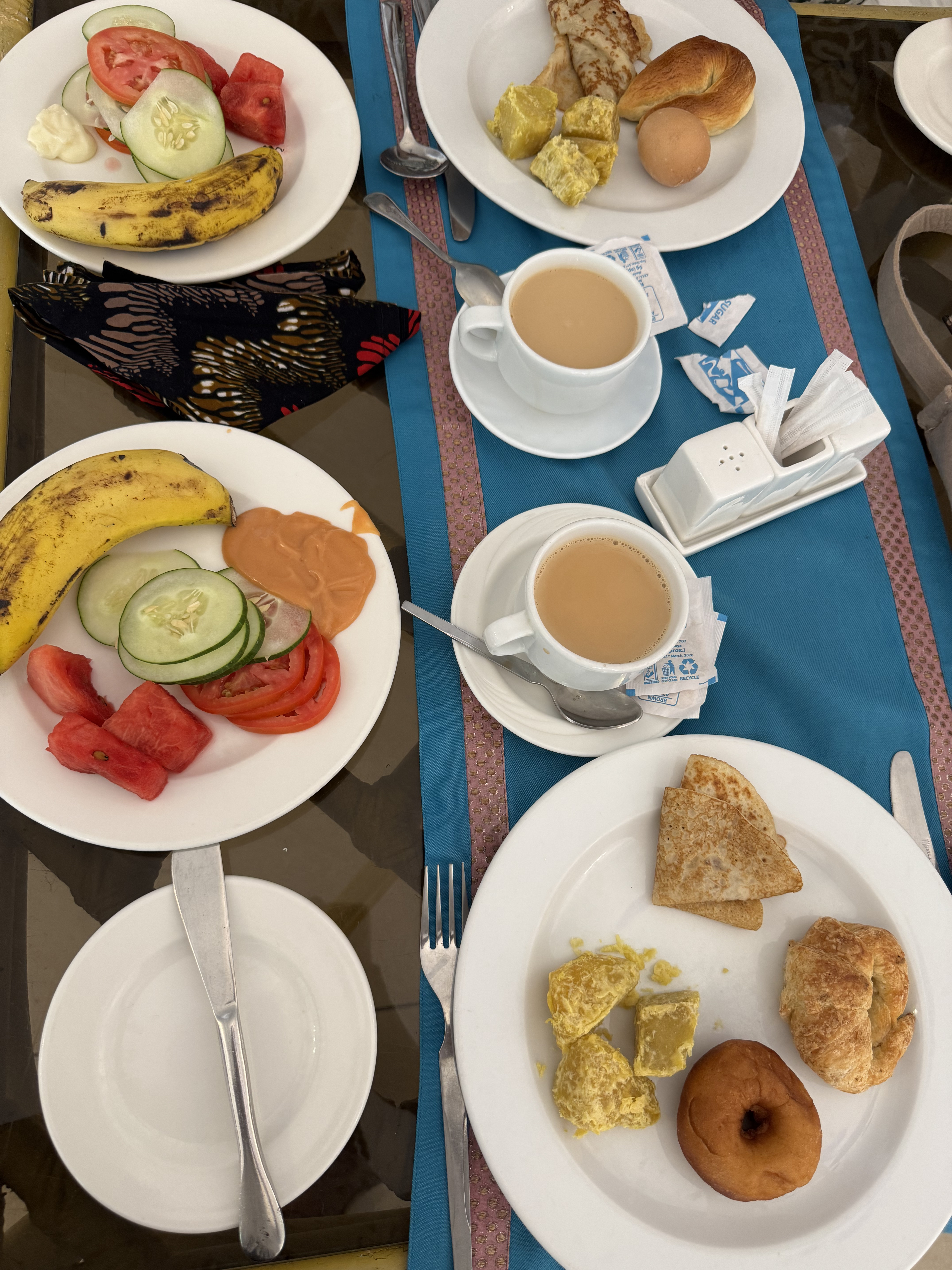 A top-down vie of a breakfast spread with plates of fruit, scrambled eggs, pastries and cups of tea on a table.
