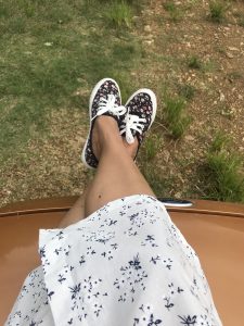 A person's legs crossed, wearing a white floral skirt and black floral sneakers, propped up on a metallic-colored surface, with grass in the background. 