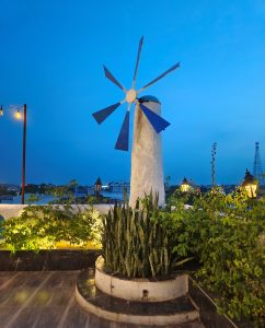 A rooftop scene illuminated by soft lighting at dusk, featuring a white cylindrical structure with a blue, multi-blade wind turbine on top. 
