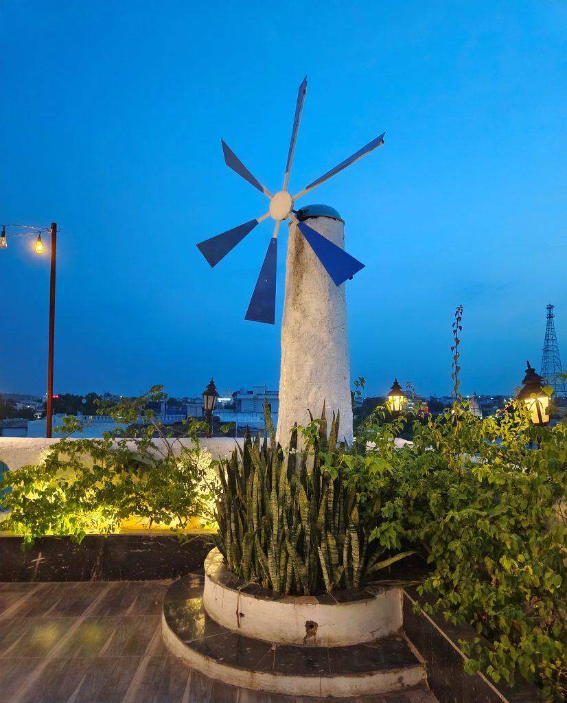 A rooftop scene illuminated by soft lighting at dusk, featuring a white cylindrical structure with a blue, multi-blade wind turbine on top.