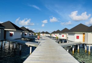 A wooden pathway extends over clear blue waters, leading to a series of overwater bungalows with sloped roofs.

