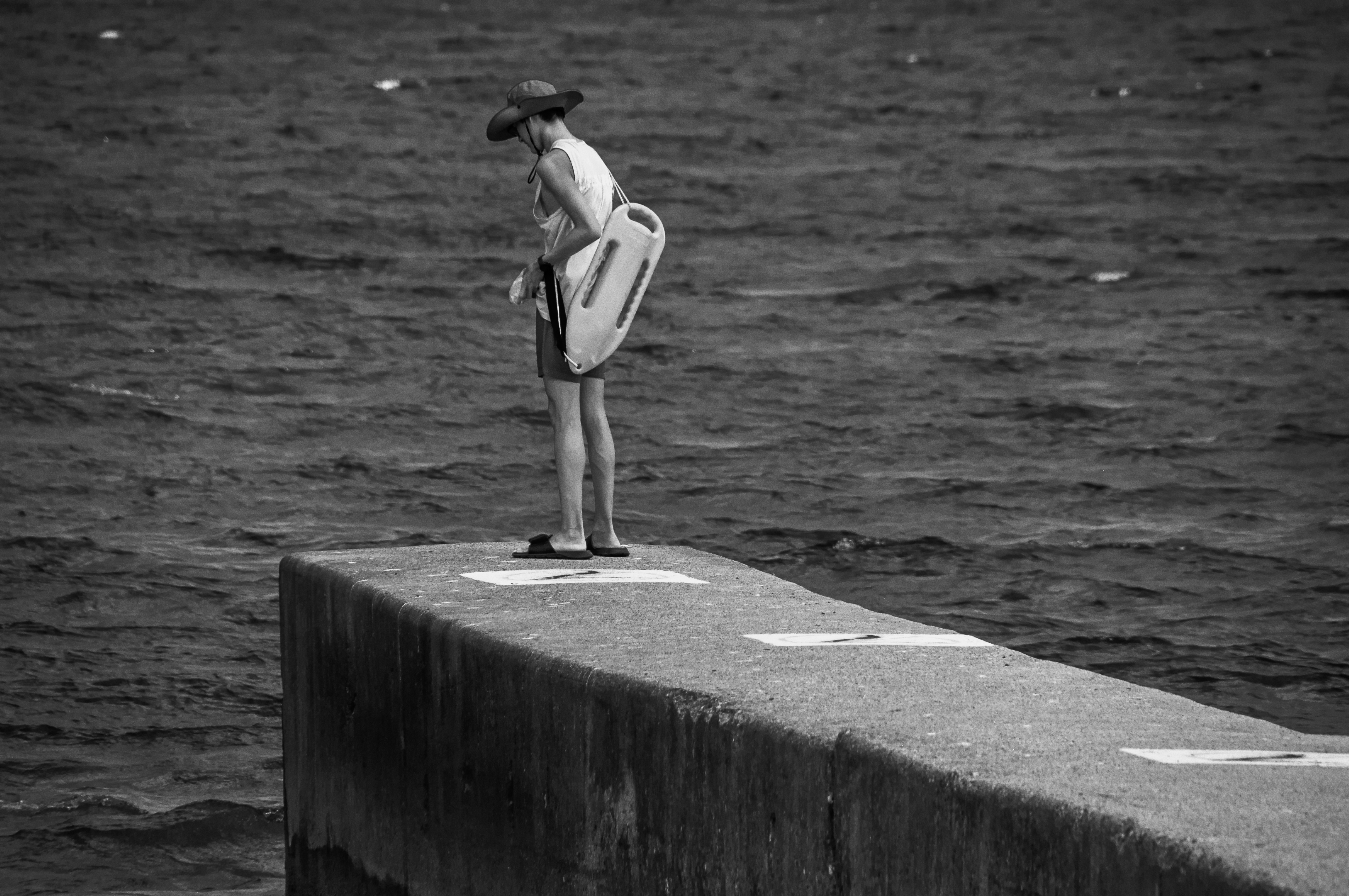 A black and white photo of a lifeguard standing at the end of a concrete pier locking down towards the water. The water has light waves with a few white caps peaking out in the distance.