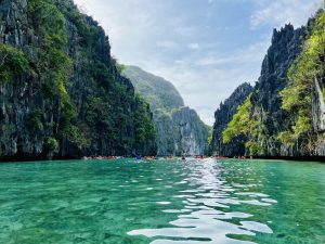 A serene turquoise lagoon surrounded by towering limestone cliffs, lush with greenery. The sky is clear blue, and kayakers explore the tranquil waters.