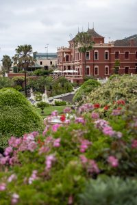 A grand red building with ornate architecture stands behind lush, manicured gardens filled with colorful flowers, palm trees, and a central fountain, under a cloudy sky. La Orotava (Tenerife-Spain)