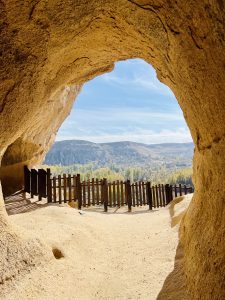 View from a cave entrance framed by rugged stone, overlooking a wooden fence and distant hilly landscape under a clear blue sky.