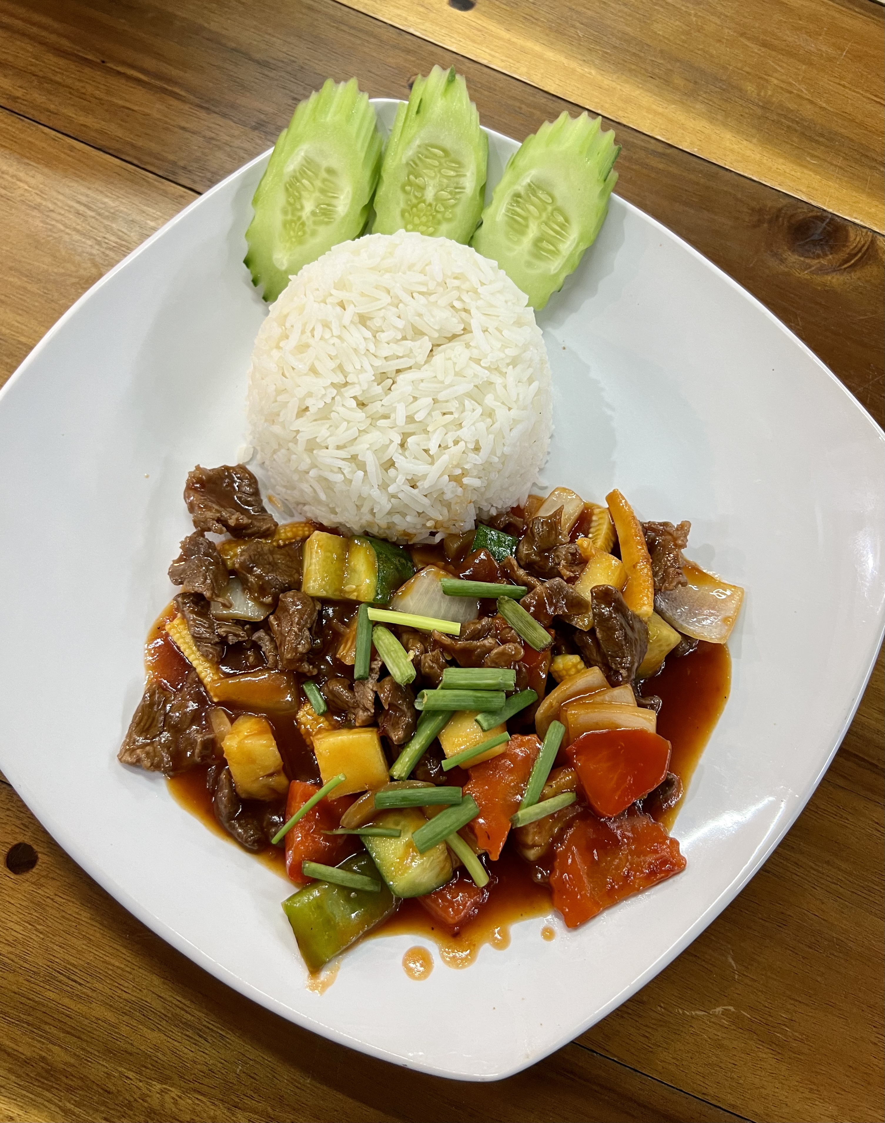 A white plate features stir-fried beef with colorful veggies, topped with green onions, alongside fluffy white rice and neatly arranged cucumber slices. Set on a wooden table.