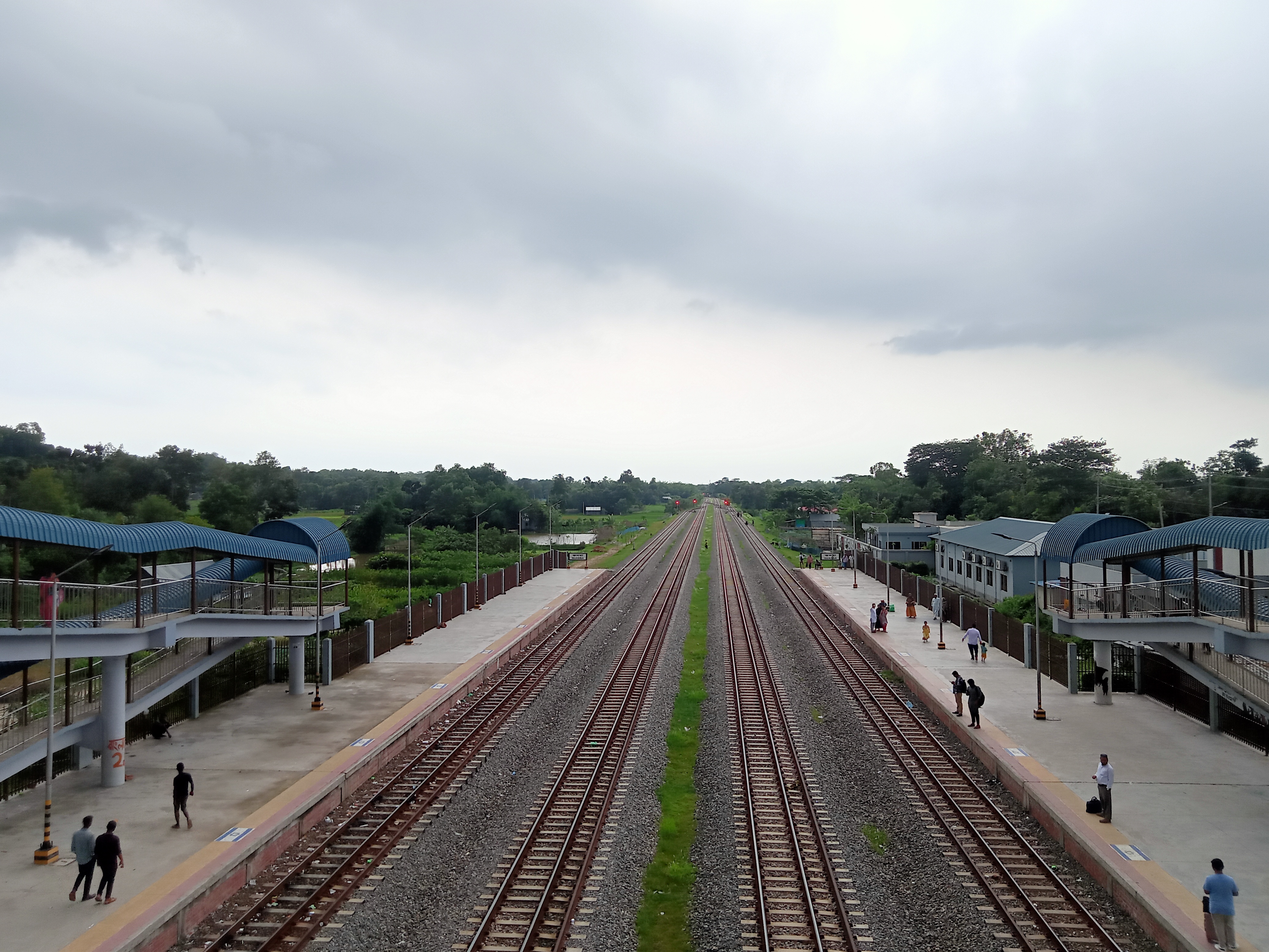 A view of a railway station featuring two parallel tracks stretching into the distance, flanked by platforms on either side. Several people are standing on the platforms, with some approaching.