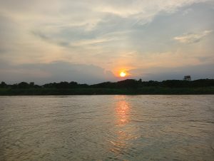 A tranquil river scene at sunset, with the sun partially hidden behind clouds, casting a warm orange glow on the water’s surface.
