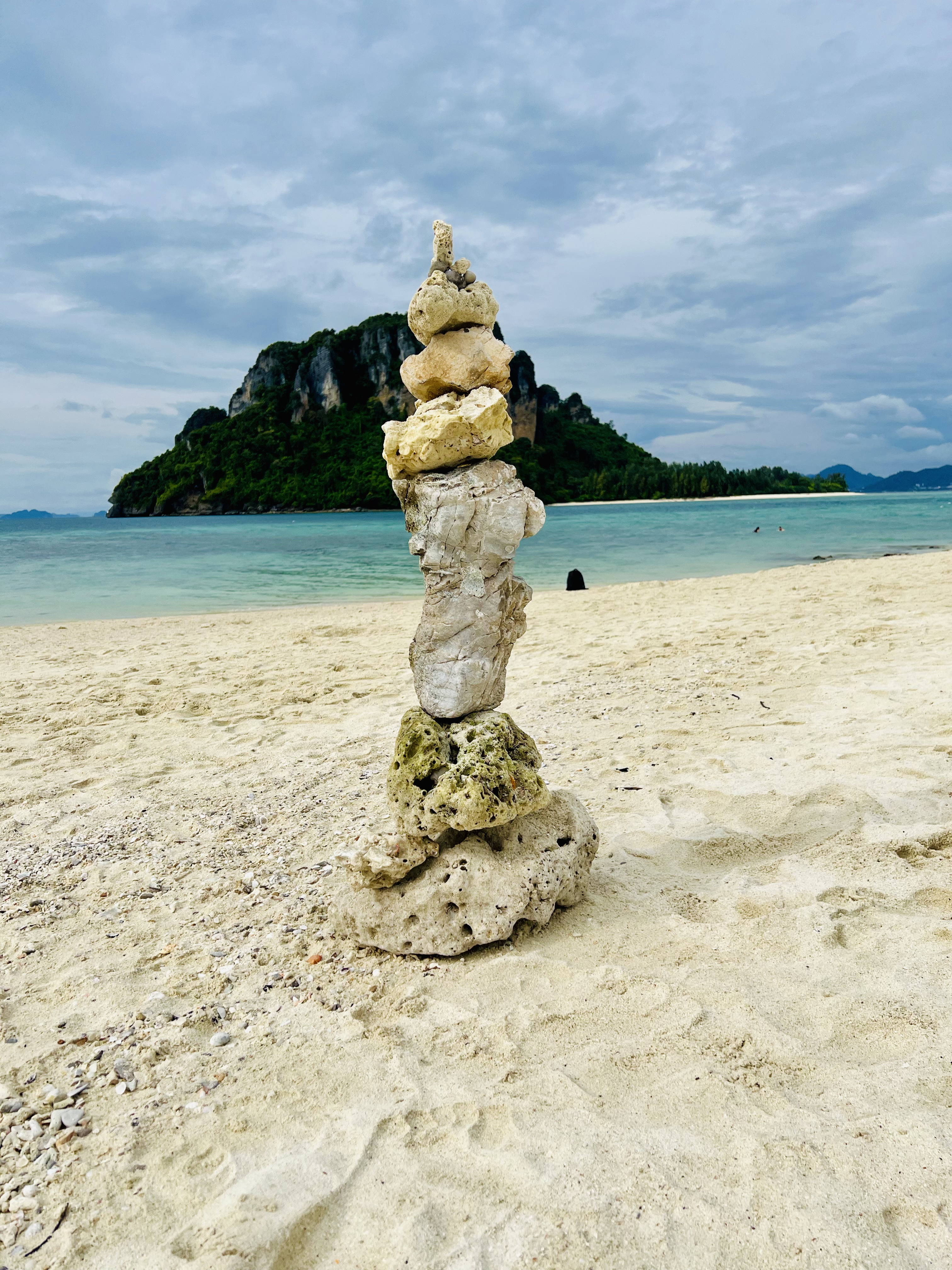 A stack of variously shaped stones balanced on a sandy beach, with a lush green island and calm turquoise waters in the background.