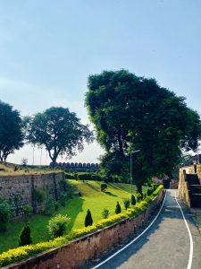 A scenic view of a pathway lined with greenery, leading towards a stone structure. Lush green trees and manicured bushes are visible alongside the path, while a blue sky fills the background. The pathway is partially shaded and curves gently, with stone walls and steps on one side leading up to a fortification in the distance. 
