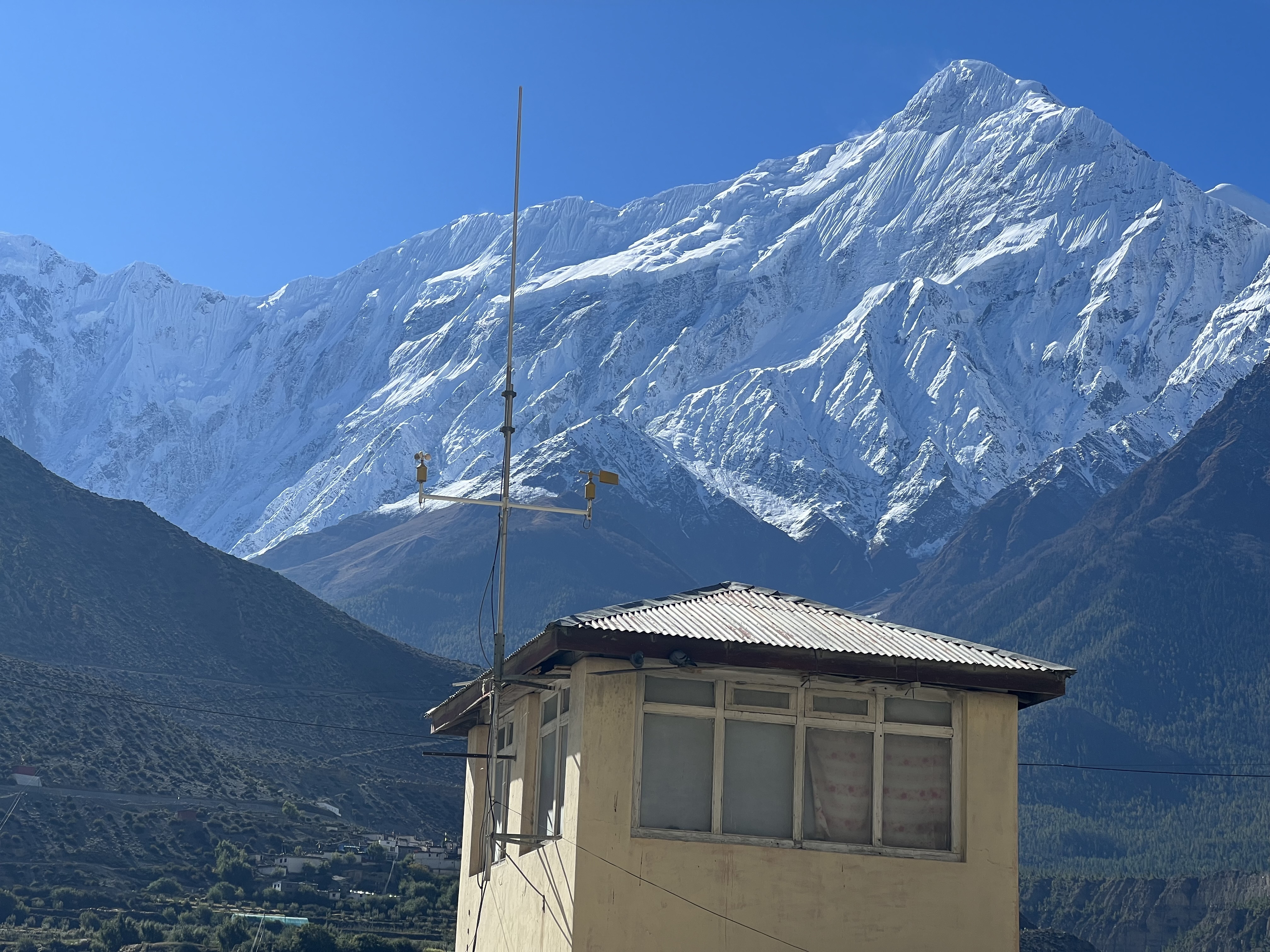 A small building with an antenna sits in front of a majestic, snow-capped mountain range under a clear blue sky.