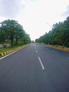 A wide, empty road stretches ahead, flanked by lush green trees on both sides. The pavement is smooth, with visible lane markings and road signs. Streetlights line the road, standing tall against the backdrop of nature.
