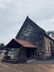 A historic stone church with a pointed roof and a large circular stained glass window, situated in a grassy area. The entrance is covered by a small awning supported by wooden columns
