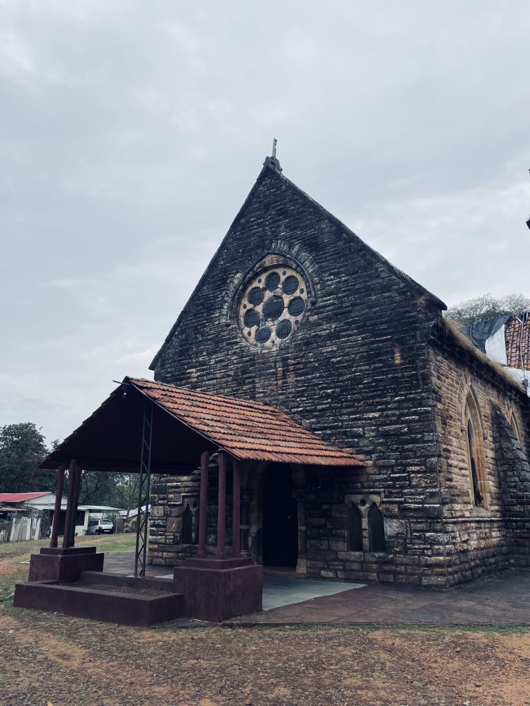 A historic stone church with a pointed roof and a large circular stained glass window, situated in a grassy area. The entrance is covered by a small awning supported by wooden columns