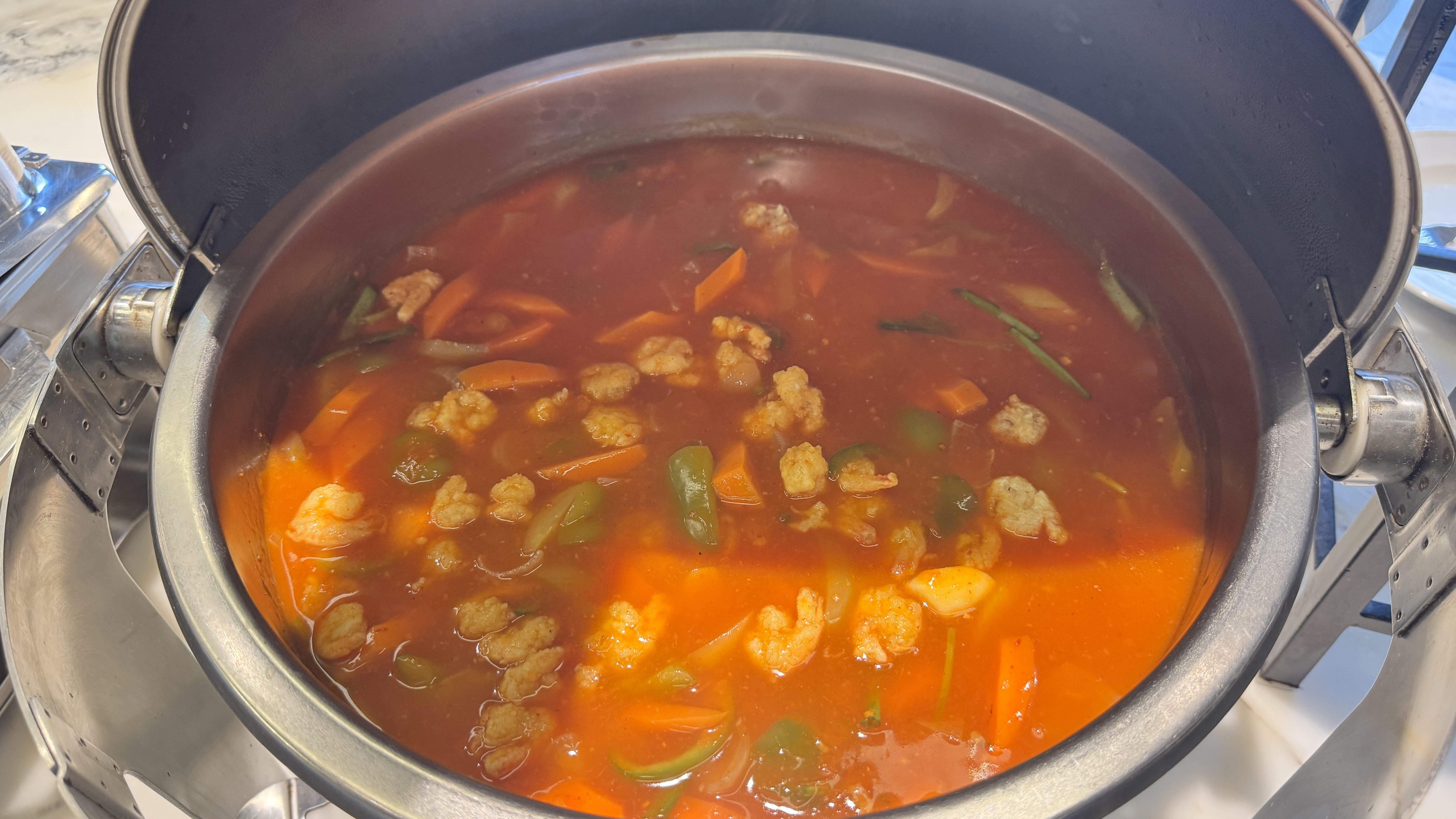 A round metal soup tureen at a buffet holds a vibrant reddish-orange broth with prawns, carrots, and green bell peppers.