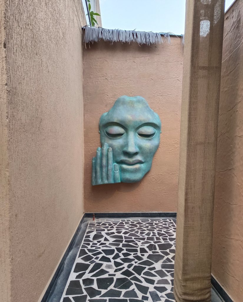 A large, turquoise-colored sculptural face is mounted on a peach-colored wall in a narrow corridor. The face features a serene expression with closed eyes and a hand resting against its cheek, suggesting contemplation.