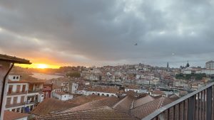A scenic view overlooking a city at sunset, with the sun casting golden hues on the horizon. The foreground features rooftops of buildings, predominantly with red-tiled roofs, while the background showcases a lively riverside city filled with various architectural styles. Vila Nova de Gaia - Porto