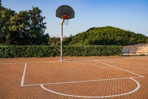 An empty outdoor basketball court with a hoop and net, set against a backdrop of green shrubs and a hillside. The court features a textured, brick-like surface and marked lines indicating the playing area 