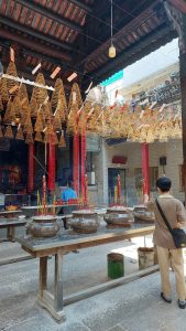 A person in a brown shirt stands in a temple courtyard, placing incense sticks into a large bronze pot while numerous hanging, spiral-shaped incense coils fill the overhead space. Captured from Vietnam.