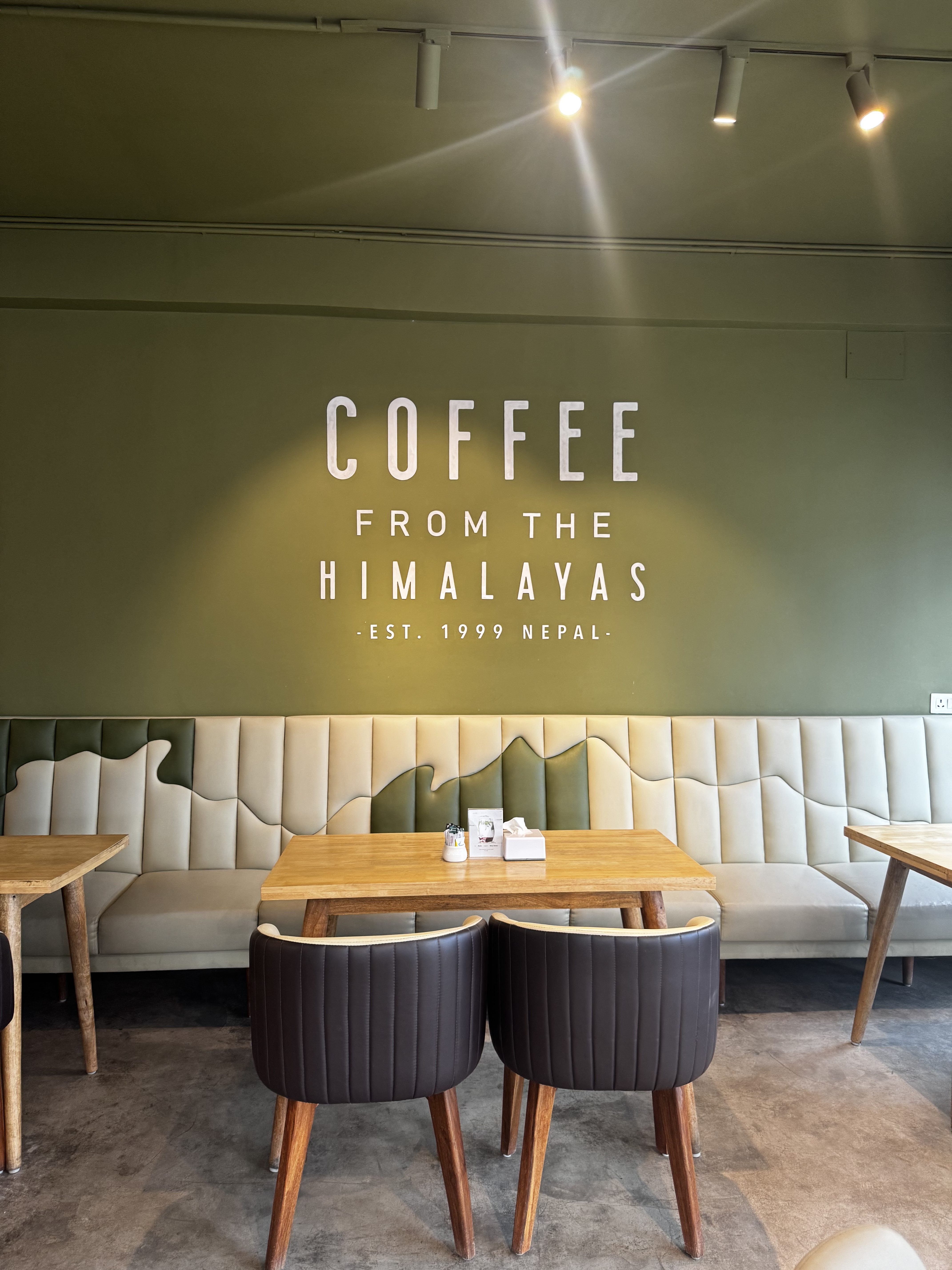 A cozy cafe interior with an olive-green wall reading “COFFEE FROM THE HIMALAYAS • EST. 1999 NEPAL.” A light bench with a mountain design lines the wall, with wooden tables and brown chairs in front.
