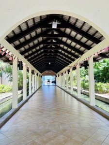 A long corridor lined with columns and a tiled floor, featuring a high, arched wooden ceiling. 