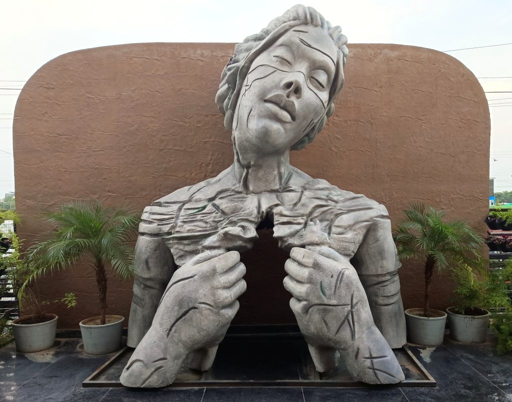 The image shows a large, grey stone sculpture of a woman whose torso is cracked and broken apart, her hands clutching the edges of the fissure. The woman’s head is tilted back, eyes closed, suggesting a state of sorrow or peace, set against a textured, earthy-brown wall with two potted palm trees framing the sculpture