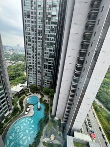 A high-angle view of a modern residential complex featuring two tall buildings flanking a lush green area. In the foreground, a curved swimming pool with a decorative leaf pattern is visible, alongside landscaped gardens and seating areas. A fountain in the center of the pool adds a serene touch. In the background, more buildings and a glimpse of a cityscape can be seen, with a road lined with vehicles to the right. The sky above is overcast, creating a muted atmosphere.
