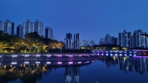 Night view of Daecheon Lake in Daecheon Park, Haeundae, Busan, with colorful lights reflecting on the water, modern sculptures in the center, and high-rise apartments in the background.