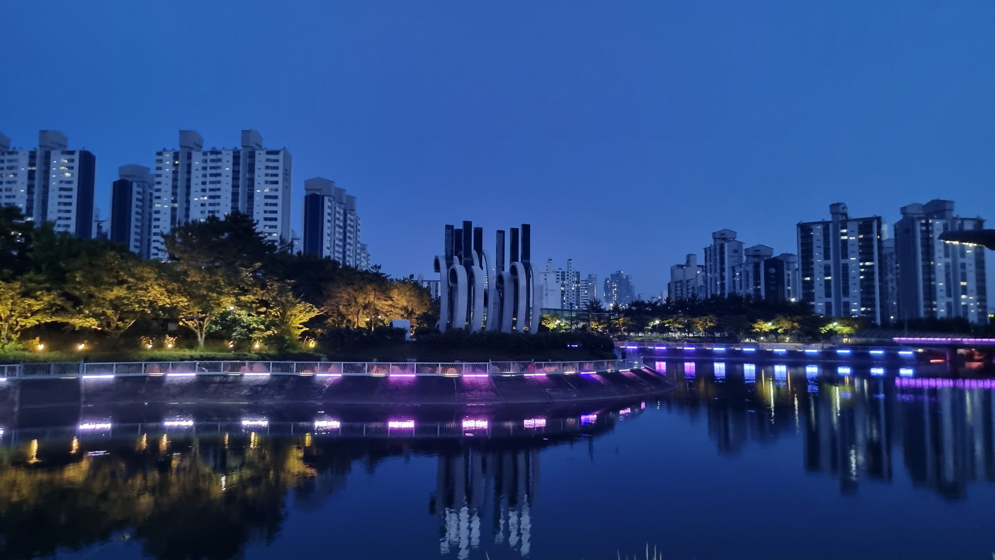 Night view of Daecheon Lake in Daecheon Park, Haeundae, Busan, with colorful lights reflecting on the water, modern sculptures in the center, and high-rise apartments in the background.
