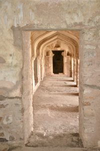 A view through a doorway into a corridor lined with arched openings, featuring rustic stone walls and a textured surface. The light creates a warm atmosphere, highlighting the intricate architectural details and the weathered surfaces of the structure. The corridor appears to lead deeper into the building, with shadows enhancing the sense of depth.