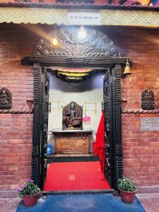 A small Hindu temple with a carved wooden doorway, red brick walls, and a brass bell. Inside, Shani Dev’s statue rests on a brick altar, with a red carpet leading in and potted plants at the entrance.
