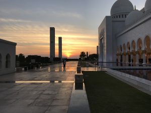 The side of a large, white mosque with a domed roof and arched windows. A person is seen walking on a tiled courtyard at sunset.