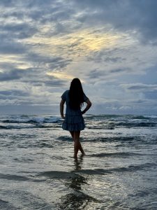 At dawn, a person stands ankle-deep in the surf, hands on hips, facing the horizon. Long dark hair and a blue patterned dress form a silhouette against layered clouds as pale gold light spreads across the reflective water.