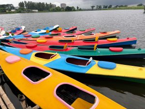 A row of colorful kayaks docked on a calm lake, with trees and buildings in the background under a cloudy sky.