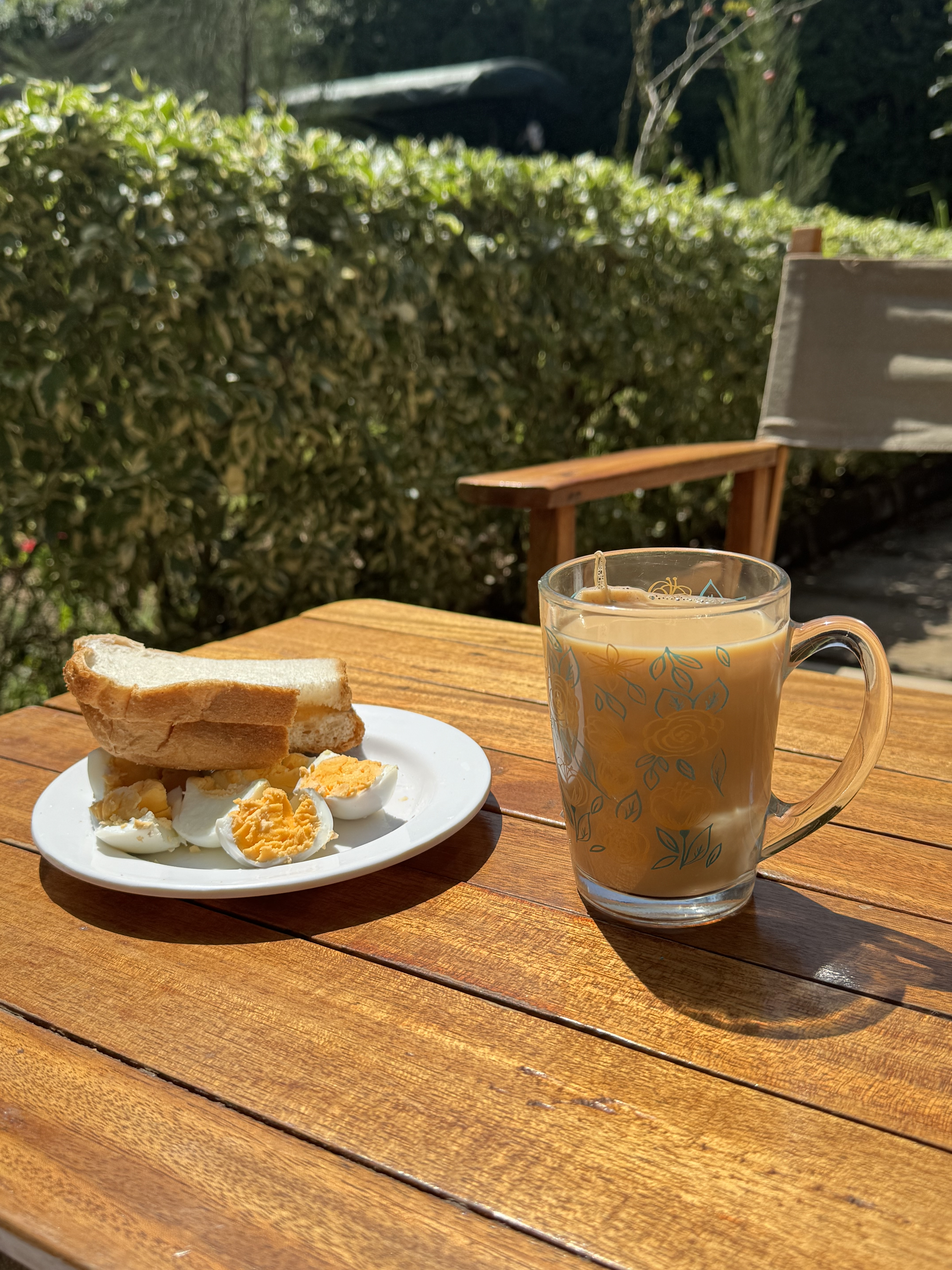 A wooden table outdoors displays a plate with sliced hard-boiled eggs and two slices of white bread. Beside the plate, a clear glass mug holds a warm cup of tea