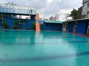 A swimming pool under a cloudy sky, with rain falling onto the water’s surface, creating ripples.
