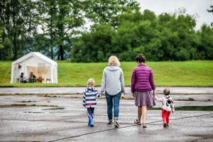Two women walking with two small children after rain on a wet pavement toward a white event tent in a green park. The group is seen from behind, holding hands, with the children wearing colorful outfits. 