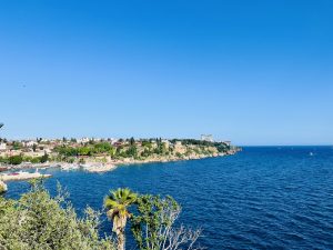 A panoramic view of a coastal city in Antalya, Turkey, featuring white buildings and a harbor full of boats, situated on a cliff overlooking the deep blue sea under a clear blue sky.