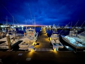 Blue hour at a marina: a long, lamp-lit dock runs to the horizon between rows of moored sportfishing boats. Calm water mirrors the lights, while a thin band of orange sunset glows beneath a deep indigo sky.