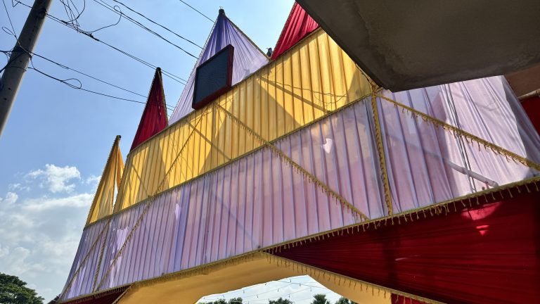 A colorful decorative structure made of draped fabric in shades of yellow, purple, and red. The fabric is arranged in a triangular pattern, creating a festive appearance. The sky is clear with some clouds, and there are power lines visible above the structure.