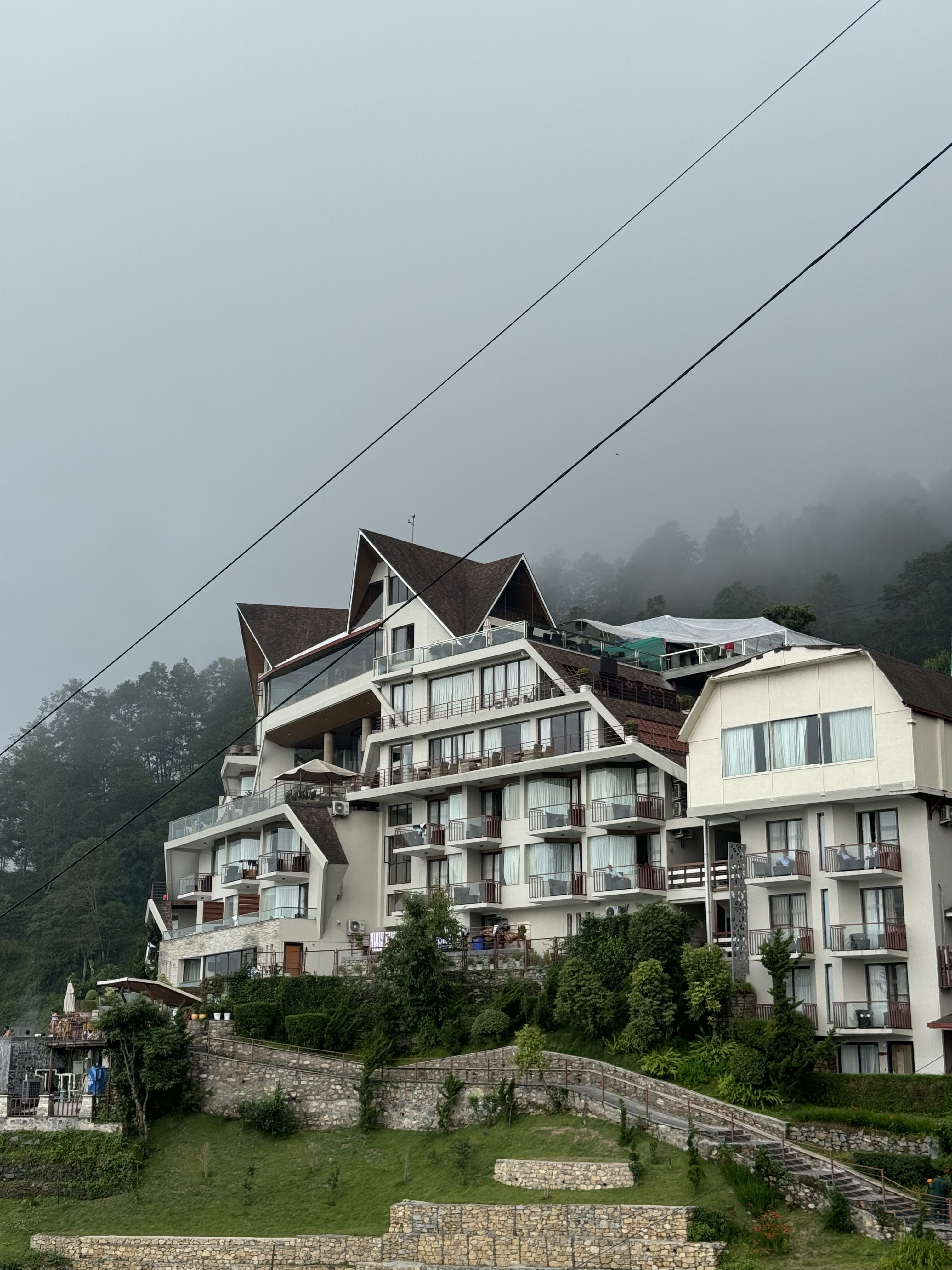 A modern multi-story building with a distinct architectural style, featuring sloping roofs and balconies. The structure is surrounded by lush greenery and stone pathways, with a hillside backdrop partially obscured by fog. The sky is overcast, creating a moody atmosphere.