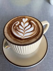 A cup of cappuccino with a detailed leaf design in the foam. The cup is a beige, ribbed ceramic with a dark brown rim, and it's on a matching saucer. 