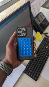 Person holding a smartphone with a blue bubble-popper-style case, in an office with a monitor, keyboard, and paperwork in the background.
